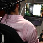 A man in a wheelchair, seen from behind, faces a laptop as he uses a Neuralink interface mounted on his chair in a home setting.