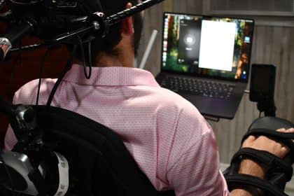A man in a wheelchair, seen from behind, faces a laptop as he uses a Neuralink interface mounted on his chair in a home setting.