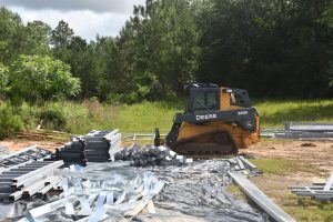 A yellow and black skid-steer loader sits near stacks of gray steel beams and metal strips at a grassy construction site.