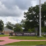 A Highland Parkway entrance sign, one-way arrow sign, sidewalk, grass and trees beneath a cloudy sky.