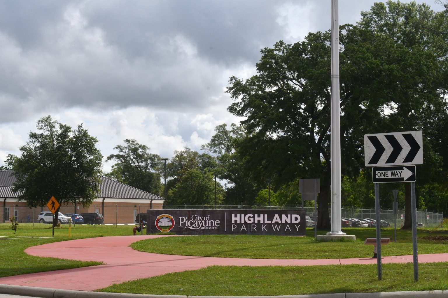 A Highland Parkway entrance sign, one-way arrow sign, sidewalk, grass and trees beneath a cloudy sky.