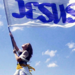 A woman stands barefoot on a grassy hill holding a large white flag with “JESUS” written in blue letters. She looks upward as the flag billows in the wind beneath a bright blue sky with scattered white clouds.