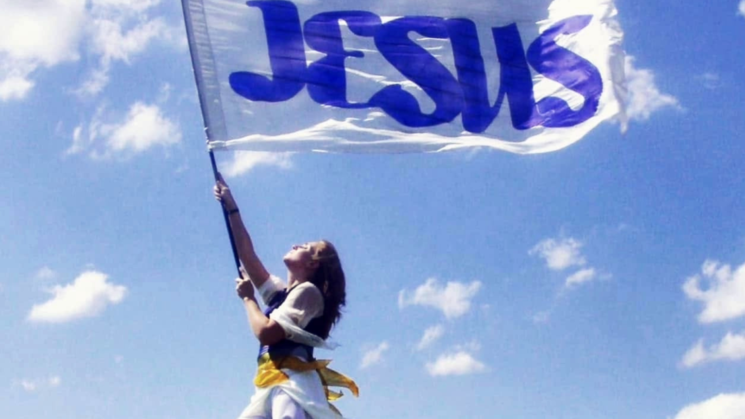A woman stands barefoot on a grassy hill holding a large white flag with “JESUS” written in blue letters. She looks upward as the flag billows in the wind beneath a bright blue sky with scattered white clouds.