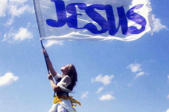 A woman stands barefoot on a grassy hill holding a large white flag with “JESUS” written in blue letters. She looks upward as the flag billows in the wind beneath a bright blue sky with scattered white clouds.