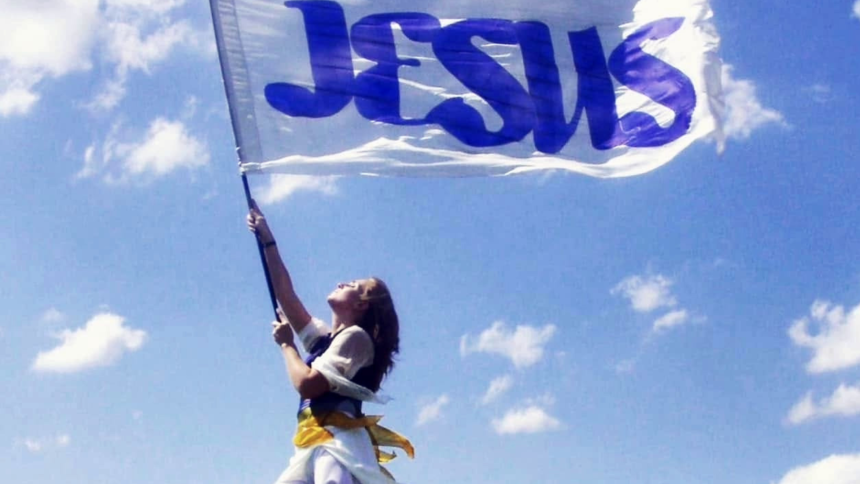 A woman stands barefoot on a grassy hill holding a large white flag with “JESUS” written in blue letters. She looks upward as the flag billows in the wind beneath a bright blue sky with scattered white clouds.
